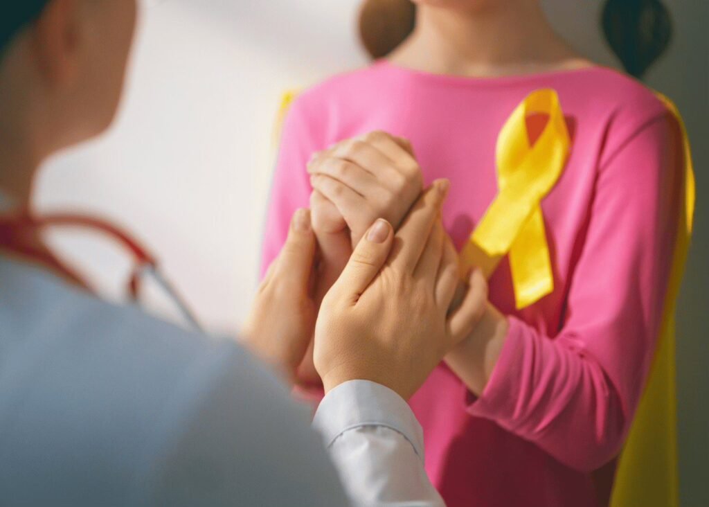 A child in a pink jumper with a yellow awareness ribbon holding a parent's hands